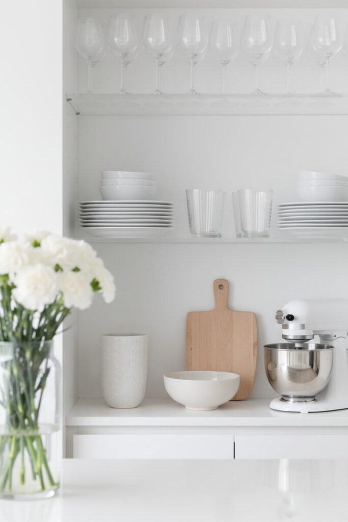 Clear acrylic shelves inside a glossy white cabinet with square plates, ribbed glasses, and a white stand mixer; soft florals blur in foreground—elegant minimalist kitchen shelf decor tips in action.