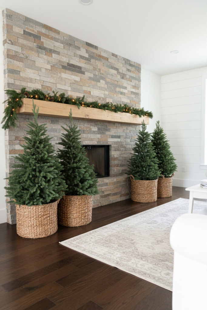 Stone-fireplace vignette with four medium artificial trees seated in richly woven wicker baskets, pine garland on mantel, and bright daylight.