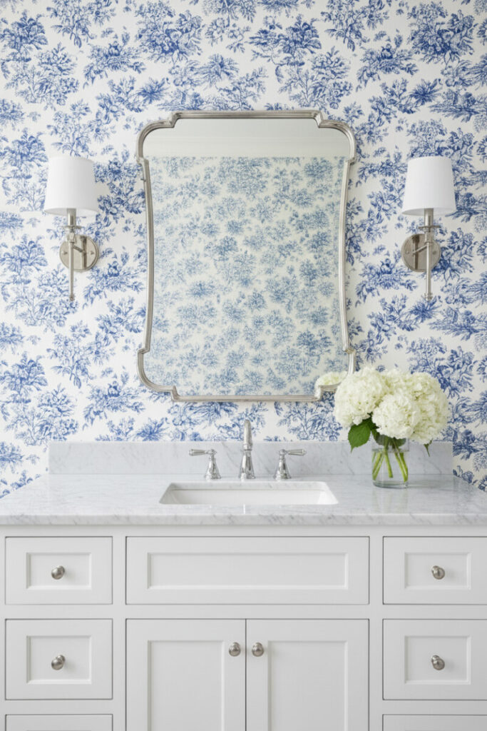Traditional vanity against intricate blue and white toile wallpaper with nickel sconces and Carrara countertop—classic detail aligned with bathroom color trends.