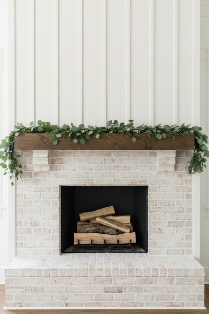White-painted brick with reclaimed wood mantel draped in eucalyptus and fir; minimalist elegance for DIY fireplace garland ideas with natural elements.