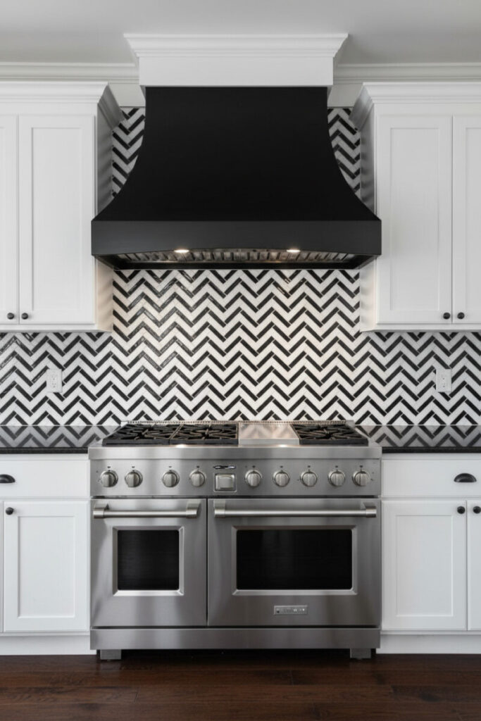 Graphic black-and-white herringbone wall with matte black hood, stainless dual-oven range, and polished black granite in dramatic task lighting.