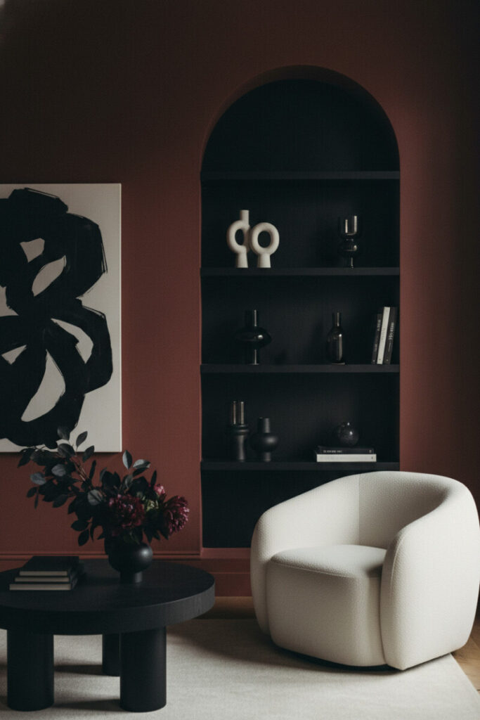 Oxblood walls with arched niche and matte-black shelving, creamy boucle chair, chunky black coffee table, and spotlit shadows—sculptural dark living room minimalism.